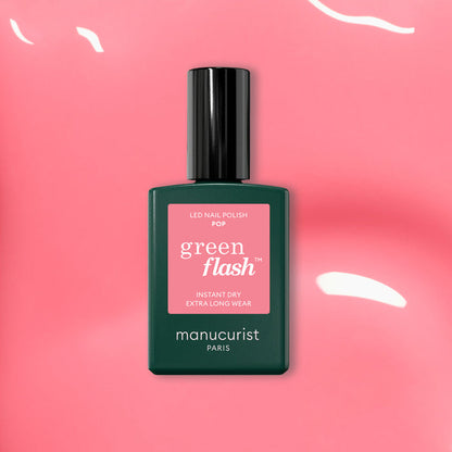A bottle of Manucurist Pop Green Flash LED nail polish, a vibrant shade with orange undertones, rests on a glossy pink surface with reflected light, its black cap and green label standing out.