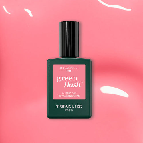 A bottle of Manucurist Pop Green Flash LED nail polish, a vibrant shade with orange undertones, rests on a glossy pink surface with reflected light, its black cap and green label standing out.