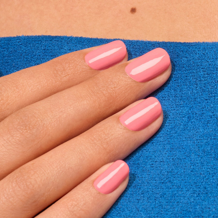 A hand with neatly manicured nails painted in Manucurist's Pop electric pink nail polish rests on a bright blue textured surface, with a small mole visible above.