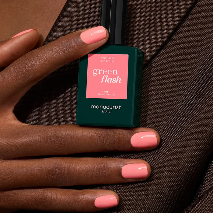 A hand with short, glossy, light pink nails holds a bottle of Manucurist Pop nail polish against a brown fabric background.
