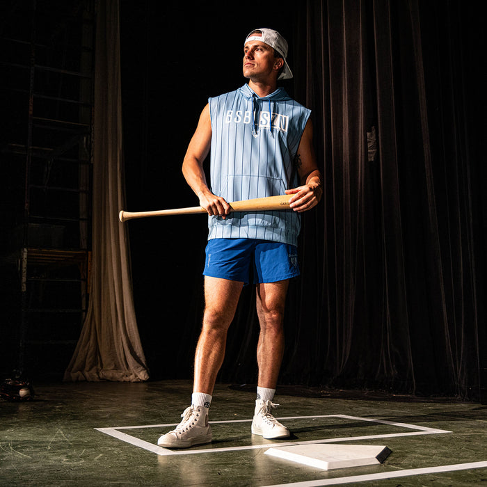 Adult male model holding a wood bat on a stage set wearing the blue Jackson Olson x BL101 Pro Series Shorts V2 paired with a matching light blue pinstripe sleeveless hoodie