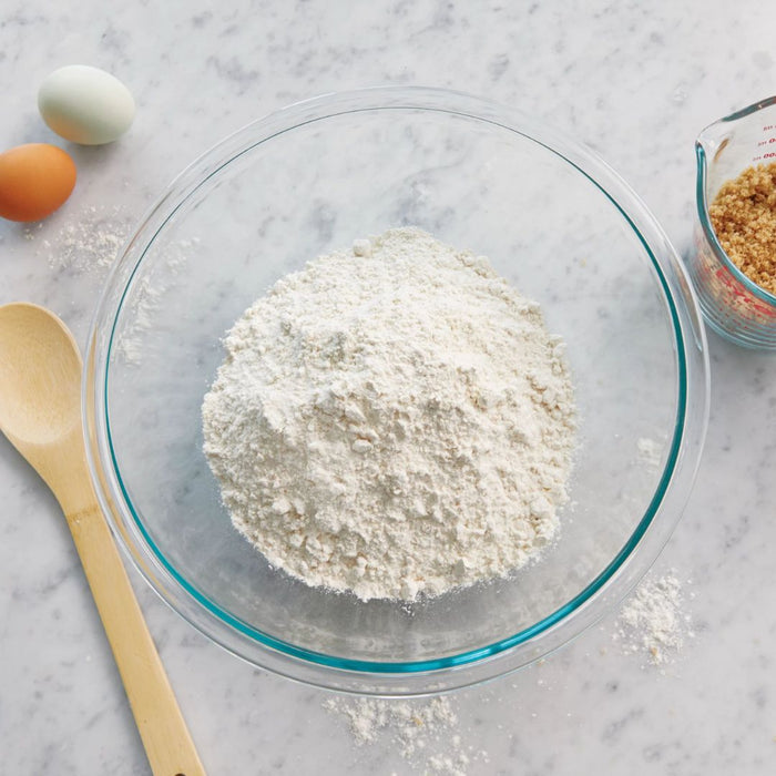 Glass bowl with flour, wooden spoon, eggs, and measuring cup on a marble surface