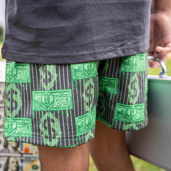 Person wearing green and gray patterned shorts with dollar bills on it, holding a suitcase