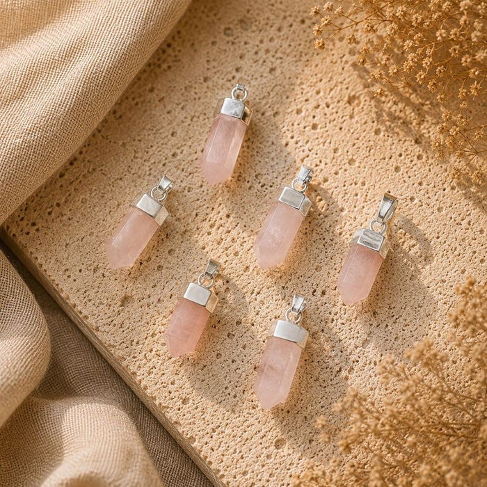 Five pink quartz crystal pendants with silver metal caps are arranged on a textured beige stone surface with draped linen and dried flowers.