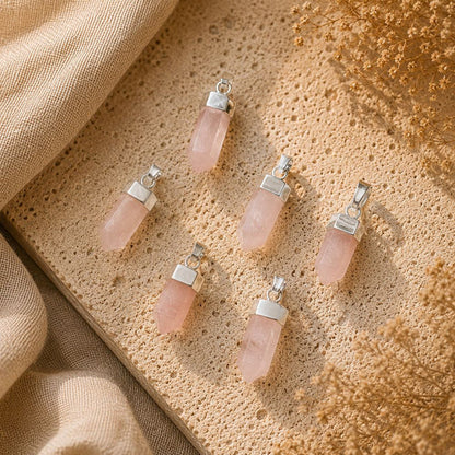 Five pink quartz crystal pendants with silver metal caps are arranged on a textured beige stone surface with draped linen and dried flowers.