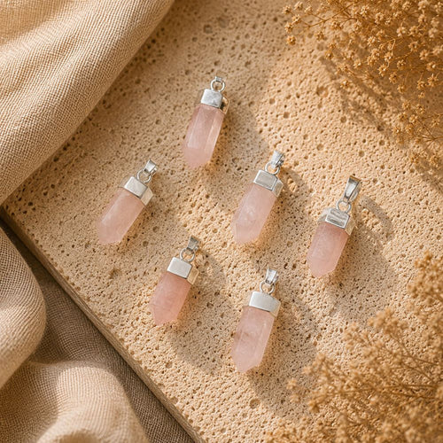 Five pink quartz crystal pendants with silver metal caps are arranged on a textured beige stone surface with draped linen and dried flowers.