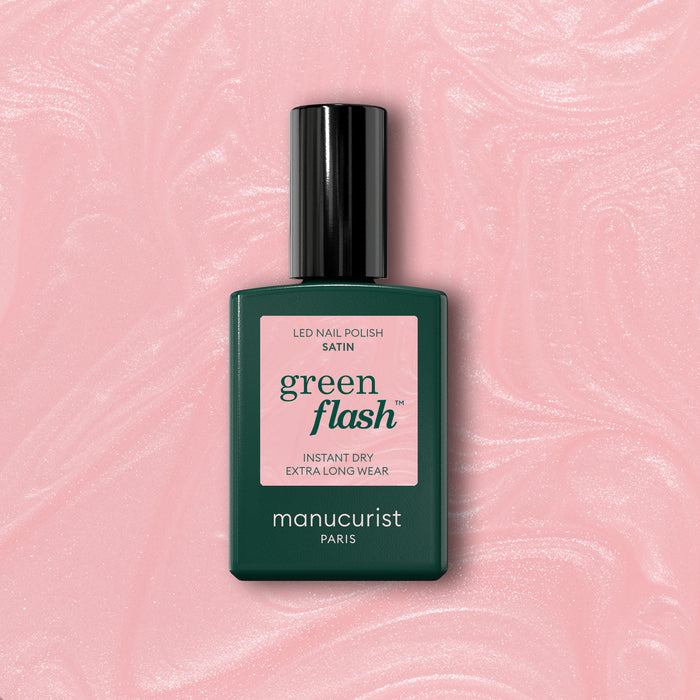 A bottle of Manucurist Satin nail polish with a pink label rests on a pale pink, shimmery background. The label reads: Satin, Green Flash, Instant Dry, Extra Long Wear.