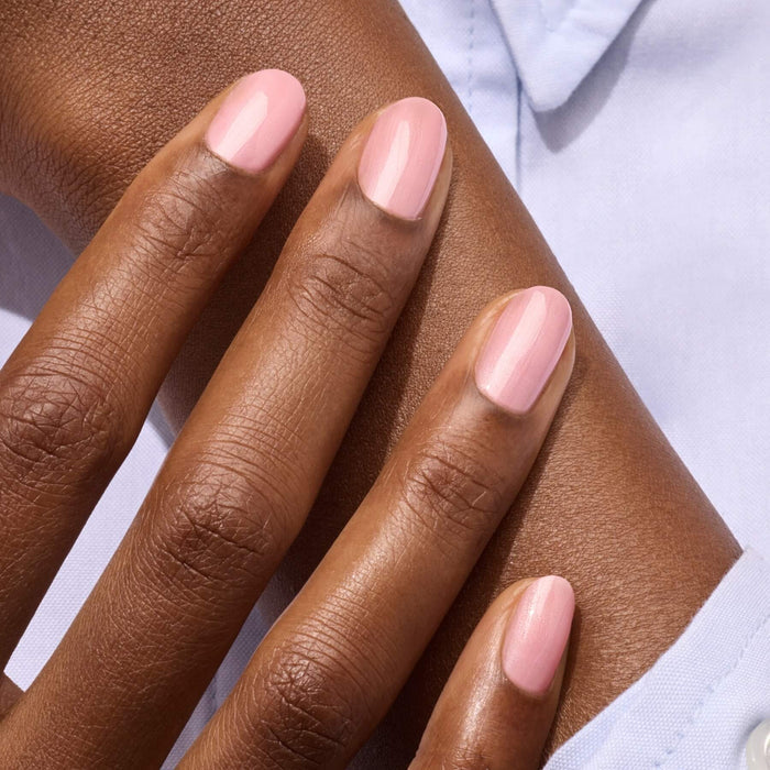 A close-up of a hand with neatly manicured nails painted in Manucurist's Satin pale pink polish, resting on a forearm. The person wears a light blue garment.