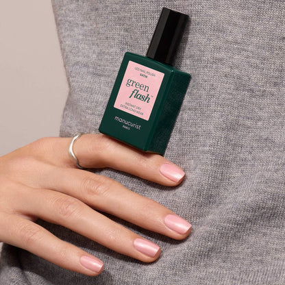 A hand with pale pink nail polish holds a bottle of Manucurist Satin nail polish against a gray sweater.