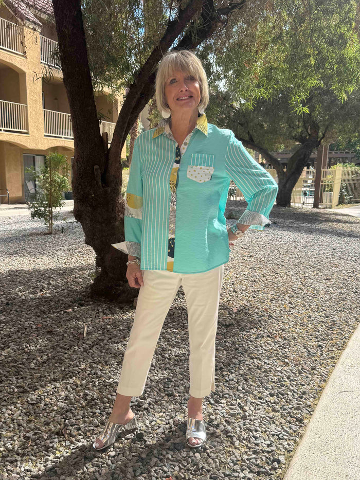Woman in a turquoise jacket and beige pants standing on a gravel path with trees and buildings in the background.