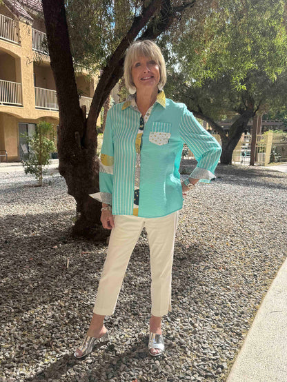 Woman in a turquoise jacket and beige pants standing on a gravel path with trees and buildings in the background.
