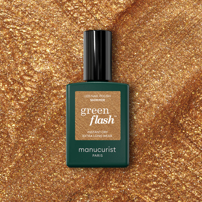 A bottle of Manucurist Shimmer gel polish sits on a sparkling gold surface that matches the cool undertone of the nail polish shade.