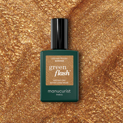 A bottle of Manucurist Shimmer gel polish sits on a sparkling gold surface that matches the cool undertone of the nail polish shade.