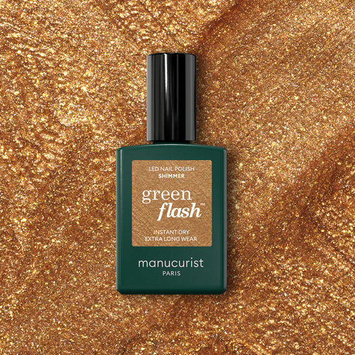 A bottle of Manucurist Shimmer gel polish sits on a sparkling gold surface that matches the cool undertone of the nail polish shade.