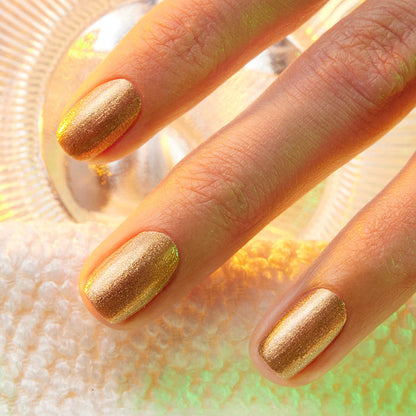 Three neatly manicured fingers showcase Manucurist’s Shimmer nail polish—a cool, shimmery gold—resting on a textured white surface with a glass object in the background.