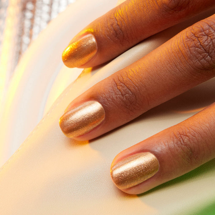 A close-up of a hand with Manucurist Shimmer gel polish in golden bronze resting on a cream surface. Warm lighting highlights the metallic sparkle on the nails.