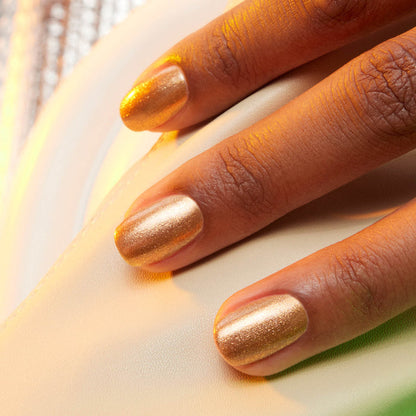 A close-up of a hand with Manucurist Shimmer gel polish in golden bronze resting on a cream surface. Warm lighting highlights the metallic sparkle on the nails.