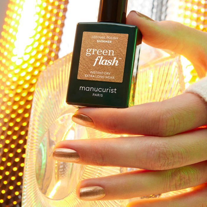A hand with shimmery gold nail polish holds a bottle of Manucurist Shimmer in front of a bright, illuminated, textured background.