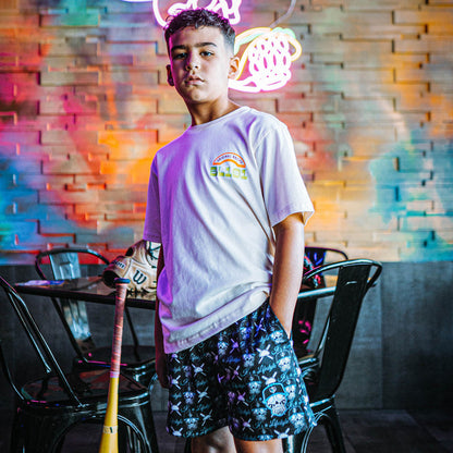 Person wearing a white t-shirt and black and blue skull shorts with colorful patterns, standing in a room with neon lights and brick walls.
