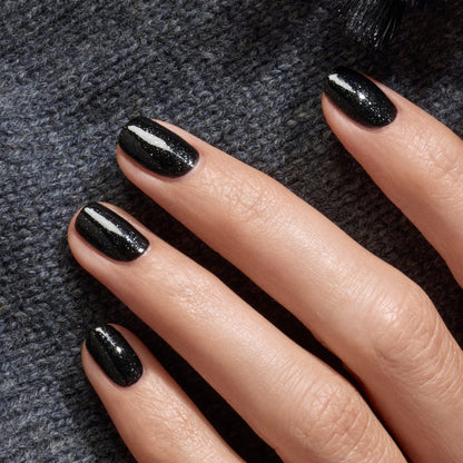 A hand with neatly manicured nails painted in Manucurist’s Sparks, a glossy black polish with a shimmering finish, rests on textured dark gray fabric.
