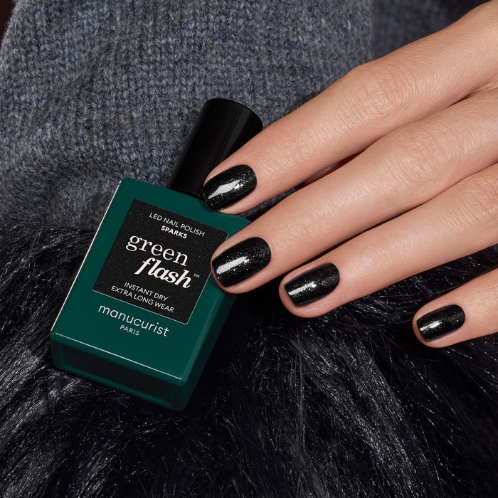 A hand with short, glossy black glitter gel polish nails rests next to a bottle of Manucurist Sparks nail polish. The background features dark, textured fabric.
