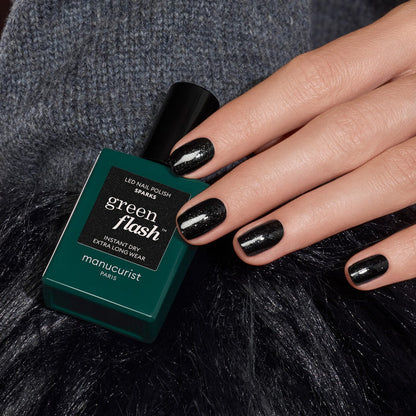 A hand with short, glossy black glitter gel polish nails rests next to a bottle of Manucurist Sparks nail polish. The background features dark, textured fabric.