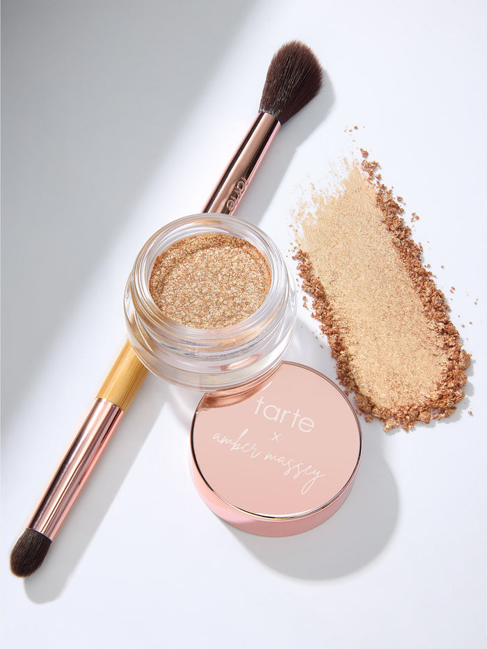 A makeup brush rests beside an open glittery gold eyeshadow pot, with a matching lid reading tarte x amber massey, on a white surface.