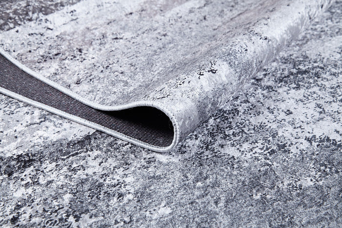 A close-up shows the corner of the Techno Flux - Washable Rug - JR1814 in gray-anthracite, slightly lifted to reveal its dark backing and edge stitching. The marbled, abstract pattern makes it ideal for high-traffic areas.