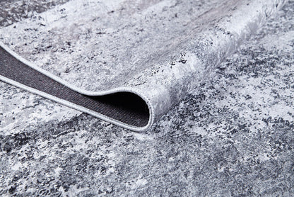 A close-up shows the corner of the Techno Flux - Washable Rug - JR1814 in gray-anthracite, slightly lifted to reveal its dark backing and edge stitching. The marbled, abstract pattern makes it ideal for high-traffic areas.