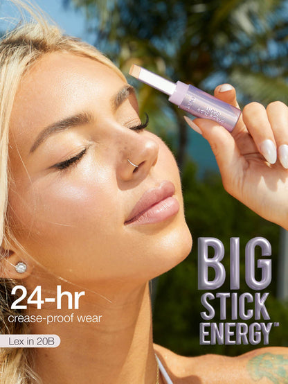 A lavender cosmetic stick is held near a person's closed eyelid; sunlit outdoor background with blurred palm trees. Text on image: 24-hr crease-proof wear; Lex in 20B; BIG STICK ENERGY