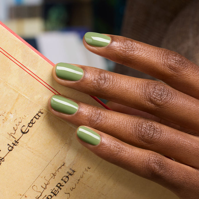 A close-up of a hand with medium brown skin and neatly manicured nails painted in Manucurist's Tuileries polish, holding a vintage yellowed paper with handwritten text.