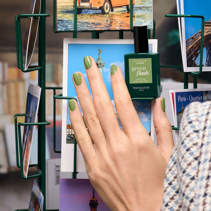 A hand with olive green gel polish holds a postcard before a rack, with Manucurist’s Tuileries Green Flash Gel Polish nearby—its color inspired by the lush beauty of Paris’s Tuileries gardens.