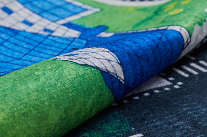 A close-up of the Urban Road - Machine-Washable Rug JRC013, a multicolor children’s play mat with illustrated roads, green spaces, blue and white geometric buildings, and a slightly curled edge.