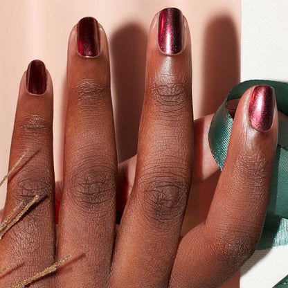 A hand with dark skin tone shows short, manicured nails painted in Manucurist's Utopia—a shiny crimson gel polish—while holding a teal ribbon against a softly blurred background.