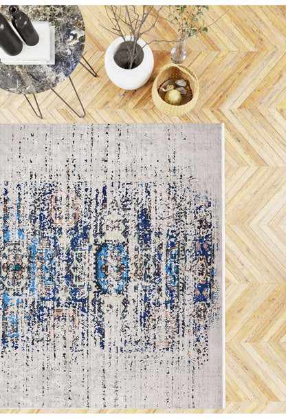 A top-down view of the Worn-Out Abstract - Washable Rug - JR339 on herringbone wood flooring, by a small table, white vase with branches, and wicker basket—ideal for high-traffic and allergy-sensitive spaces.